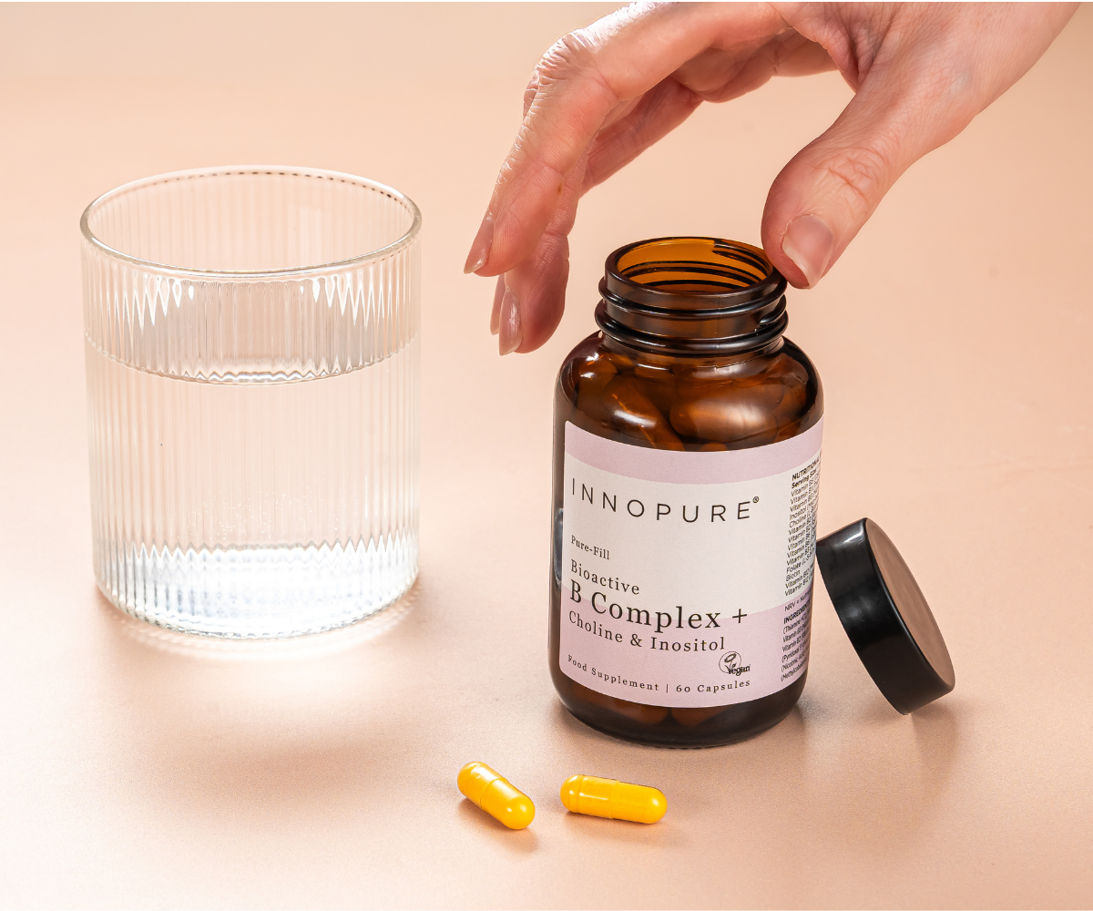 Open jar of Innopure Bioactive B Complex + Choline & Inositol with a hand reaching in, beside a glass of water and two yellow capsules