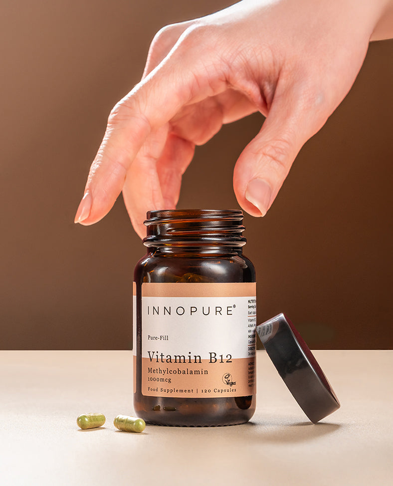 Innopure Vitamin B12 Methylcobalamin 1000mcg amber glass jar, lid off, with a hand reaching in and two green capsules beside it