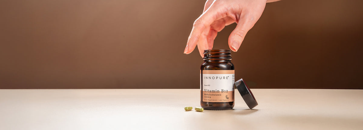 Innopure Vitamin B12 Methylcobalamin 1000mcg amber glass jar, lid off, with a hand reaching in and two green capsules beside it