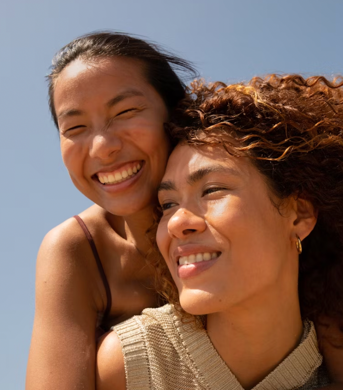 Two women laughing together with their heads close, in bright sunshine against a blue sky